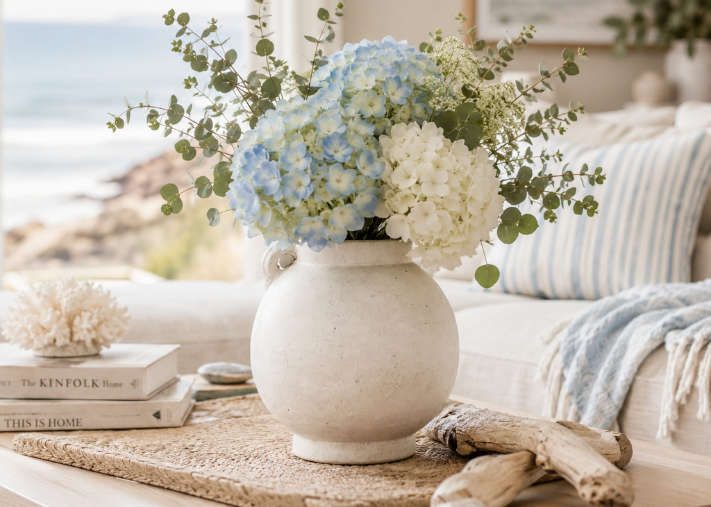 A ceramic vase filled with light blue and white hydrangeas and green eucalyptus stems, styled on a table in a bright coastal living room with a window, pillows, and books.