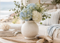 A ceramic vase filled with light blue and white hydrangeas and green eucalyptus stems, styled on a table in a bright coastal living room with a window, pillows, and books.