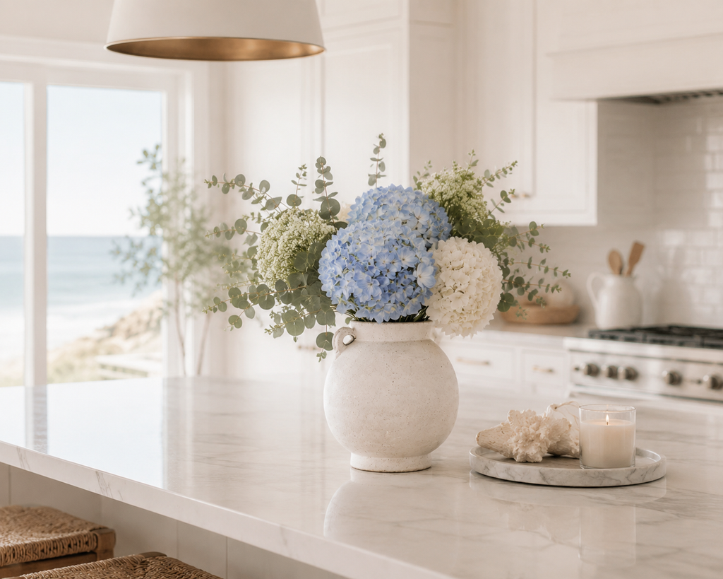 A decorative ceramic vase filled with blue and white hydrangeas and green foliage, styled on a white marble kitchen island with a candle and seashells nearby, overlooking the ocean.